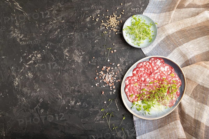 Slices of smoked cervelat salami sausage with spinach microgreen, salt and pepper on black concrete background and beige textile. Top view, flat lay, copy space