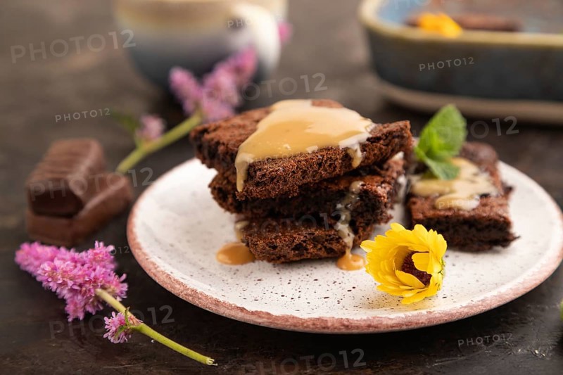 Chocolate brownie with caramel sauce with a cup of coffee on black concrete background. side view, close up, selective focus