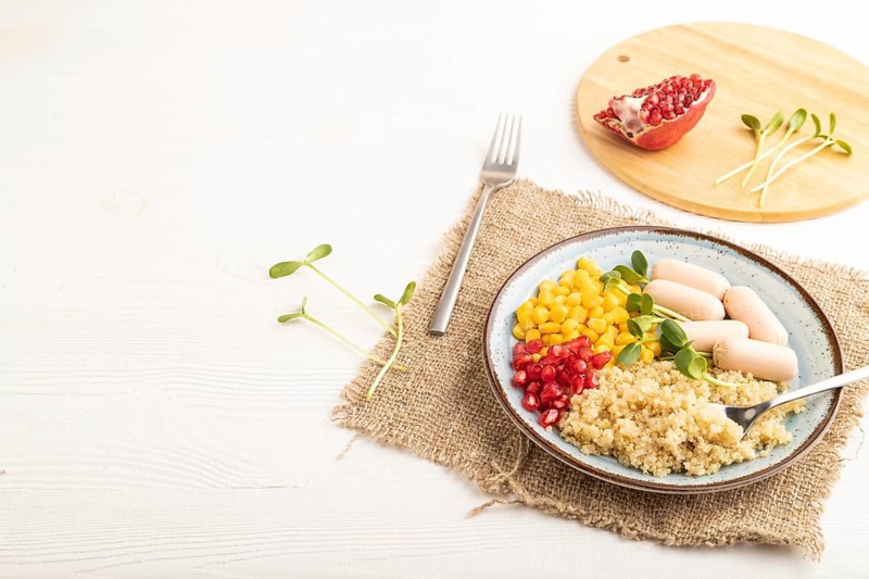 Mixed quinoa porridge, sweet corn, pomegranate seeds and small sausages on white wooden background. Side view, copy space. Food for children, healthy food concept