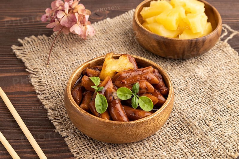 Tteokbokki or Topokki, fried rice cake stick, popular Korean street food with spicy jjajang sauce and pineapple on brown wooden background and linen textile. Side view