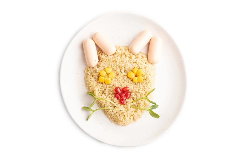 Funny mixed quinoa porridge, sweet corn, pomegranate seeds and small sausages in form of cat face isolated on white background. Top view, flat lay, close up. Food for children, healthy food concept