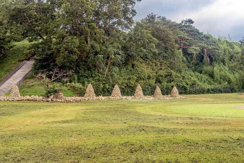 Row of cairn (towers of stack stones) in front of treeline with stairs up side of hill on left in South Korea