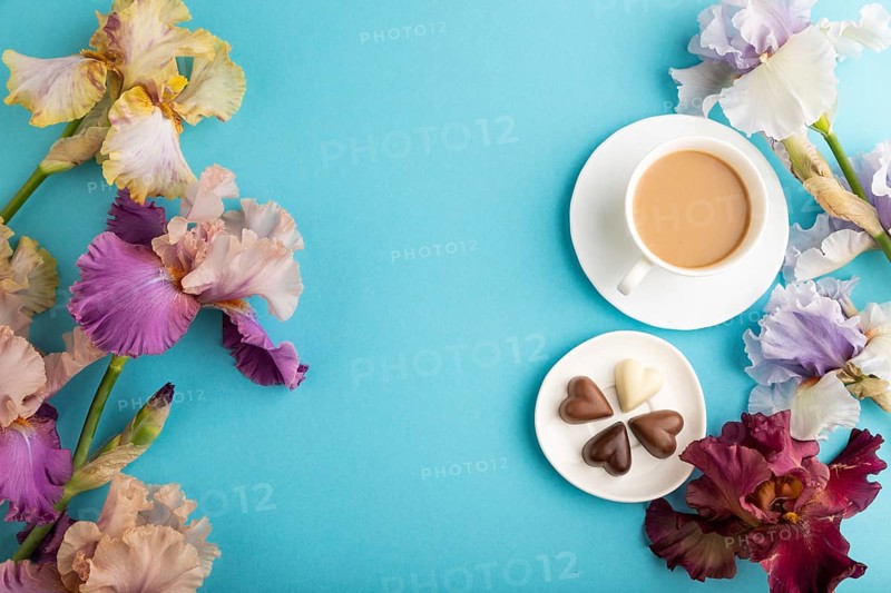 Cup of cioffee with chocolate candies, lilac and purple iris flowers on blue pastel background. top view, flat lay, copy space, still life. Breakfast, morning, spring concept