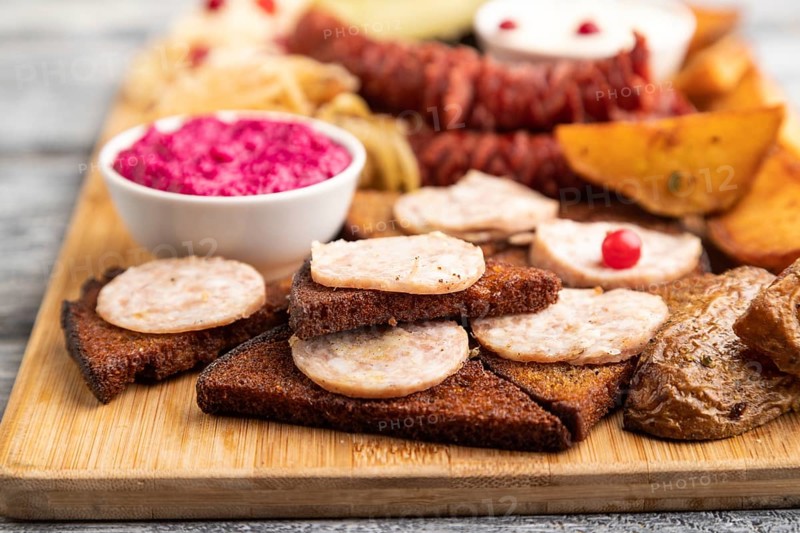 Set of snacks: sausages, toast, sauerkraut, marinated onion and cucumber, baked potato on a cutting board on a gray wooden background. Side view, close up, selective focus