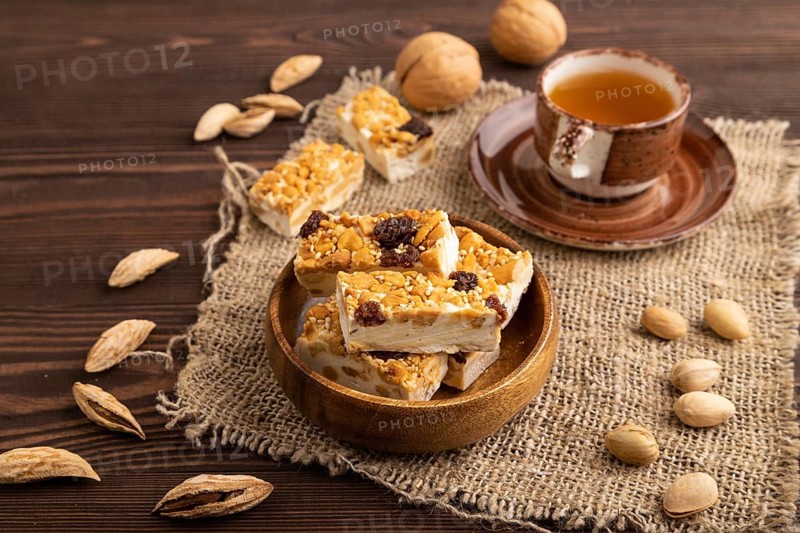 Traditional candy nougat with nuts and sesame with cup of green tea on brown wooden background and linen textile. side view, close up