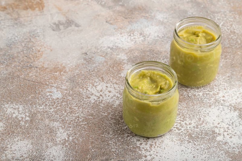 Baby puree with vegetable mix, broccoli, tomatoes, cucumber, avocado infant formula in glass jar on brown concrete background. Side view, copy space, artificial feeding concept