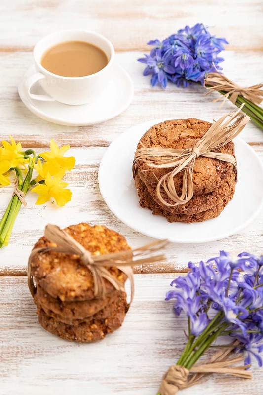 Oatmeal cookies with spring snowdrop flowers bluebells, narcissus and cup of coffee on white wooden background. side view, close up, still life. Breakfast, morning, spring concept