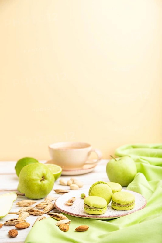 Green macarons or macaroons cakes with cup of coffee on a white and orange background and green linen textile. Side view, copy space, selective focus