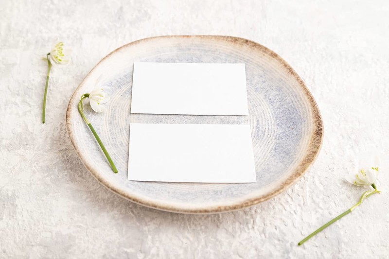 White paper invitation card, mockup with galanthus snowdrop flowers on ceramic plate and gray concrete background. Blank, side view, still life, copy space, wedding invitation