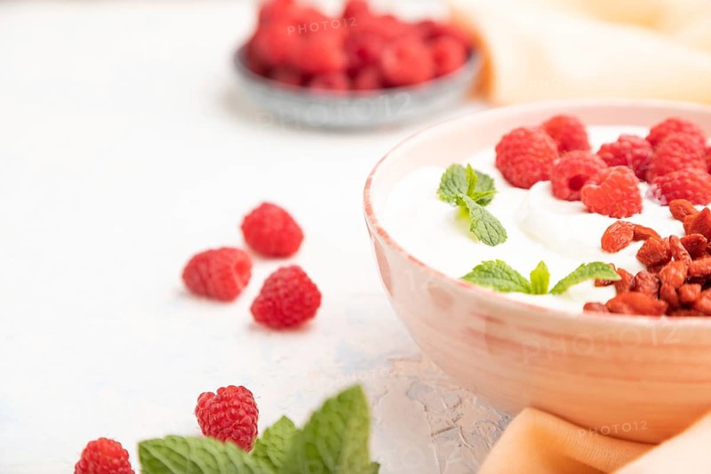 Yogurt with raspberry and goji berries in ceramic bowl on white concrete background and orange linen textile. Side view, close up, selective focus