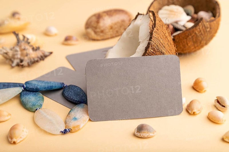 Brown paper business card with coconut, seashells, pebbles, beads on orange pastel background. Side view, copy space. Tropical, healthy food, vacation, holidays concept