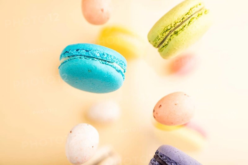 Multicolored flying macaroons and chocolate eggs frozen in the air on blurred beige background. top view, flat lay, close up. Breakfast, morning, concept