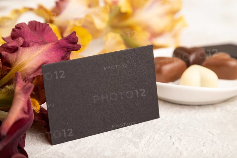 Black business card with chocolate candies and iris flowers on gray concrete background. side view, copy space, still life. Breakfast, morning, spring concept