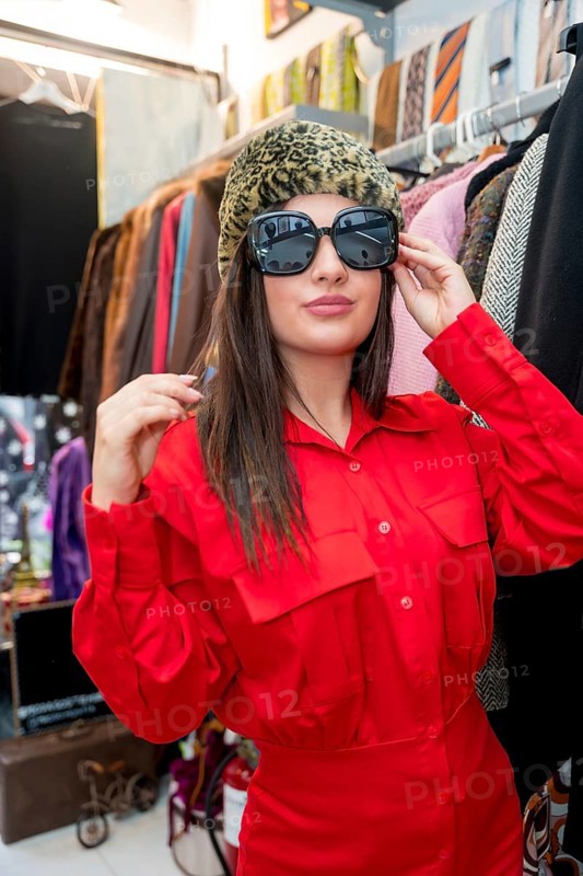 Vertical portrait of a stylish woman trying on a hat and sunglasses in a retro clothes store
