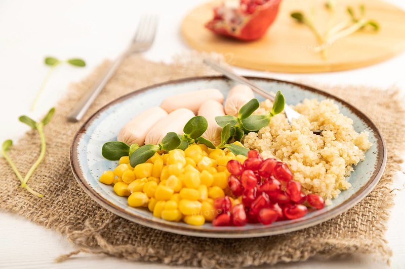 Mixed quinoa porridge, sweet corn, pomegranate seeds and small sausages on white wooden background. Side view, close up, selective focus. Food for children, healthy food concept