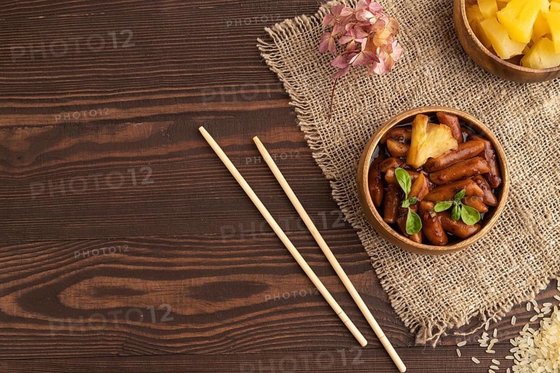 Tteokbokki or Topokki, fried rice cake stick, popular Korean street food with spicy jjajang sauce and pineapple on brown wooden background and linen textile. Top view, flat lay, copy space