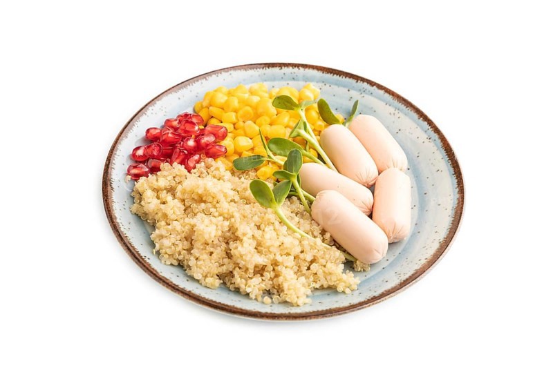 Mixed quinoa porridge, sweet corn, pomegranate seeds and small sausages isolated on white background. Side view, close up. Food for children, healthy food concept