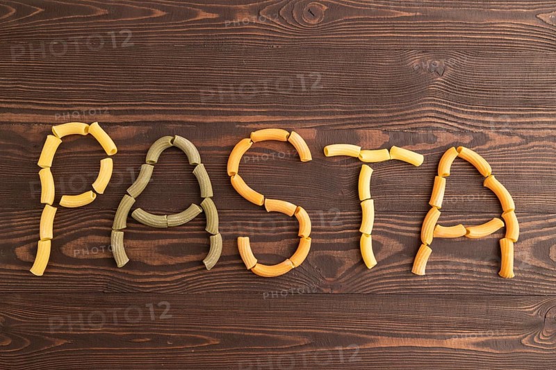Inscription pasta, Rigatoni colored raw pasta with tomato, eggs, spices, herbs on brown wooden background. Top view, flat lay, copy space