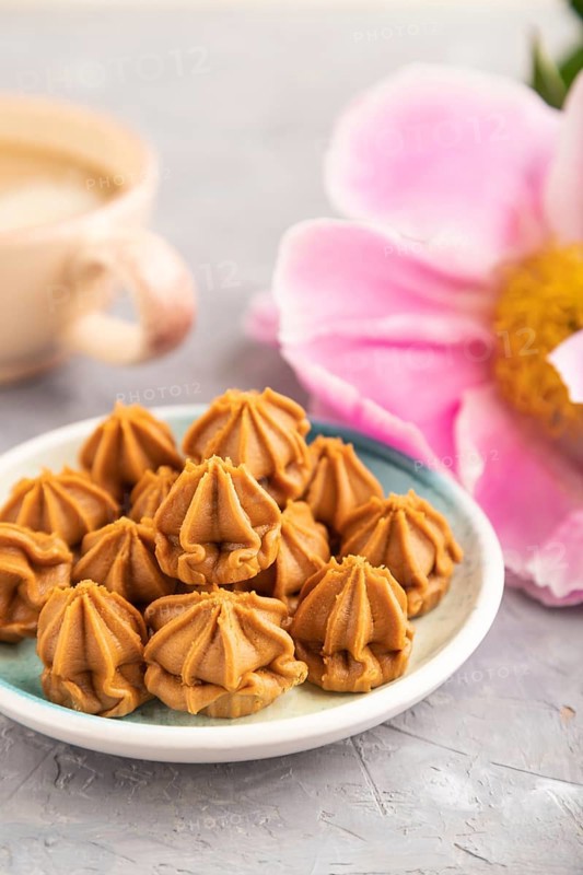 Homemade soft caramel fudge candies on blue plate and cup of coffee on gray concrete background, peony flower decoration. side view, close up, selective focus