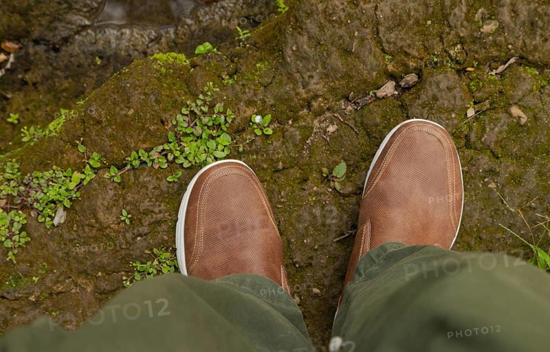 Man feets in hiking leather shoes in outdoors. Top View of shoes on rocky srones near river waterfall, travelling concept