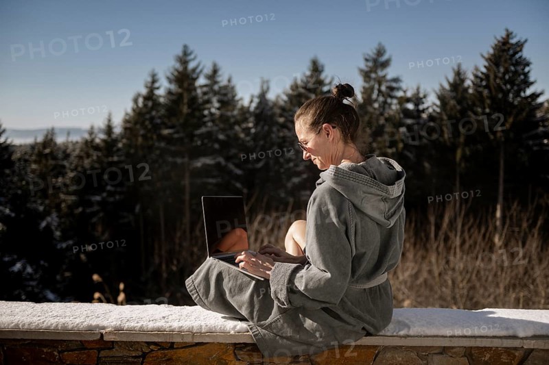 Woman in a bathrobe sits on a snowy stone wall in nature and works on her laptop