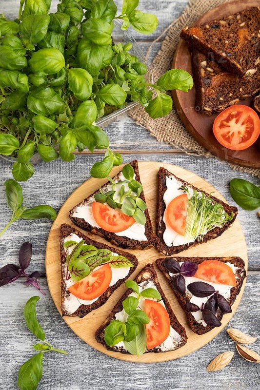Grain rye bread sandwiches with cream cheese, tomatoes and basil microgreen on gray wooden background and linen textile. top view, flat lay, close up
