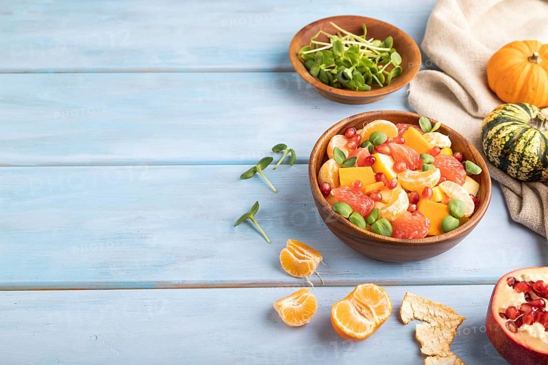Vegetarian fruit salad of pumpkin, tangerine, pomegranate, grapefruit, sunflower microgreen sprouts on blue wooden background and linen textile. Side view, copy space