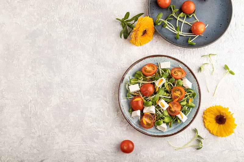 Vegetarian vegetables salad of tomatoes, marigold petals, microgreen sprouts, feta cheese on gray concrete background. Top view, flat lay, copy space