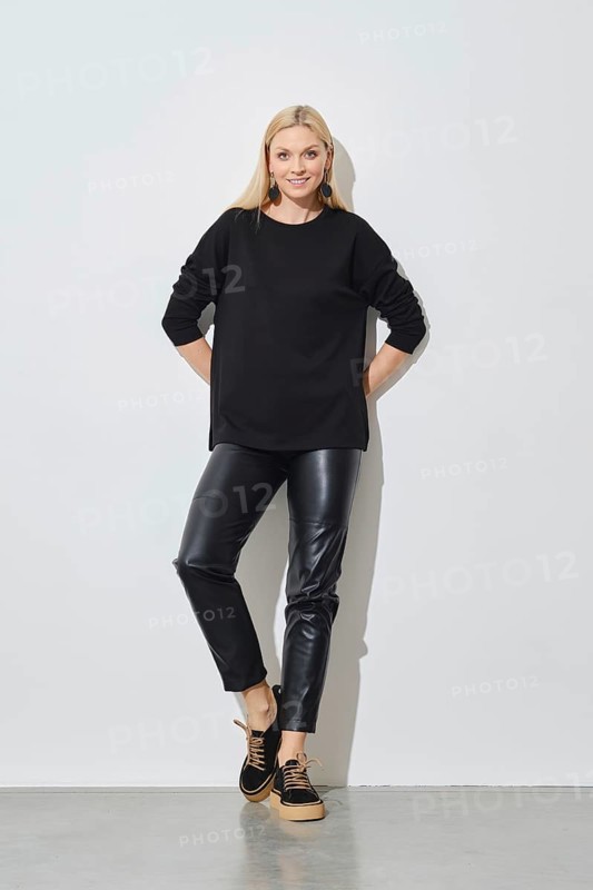 Smiling woman in black pullover and leather trousers leaning against white wall
