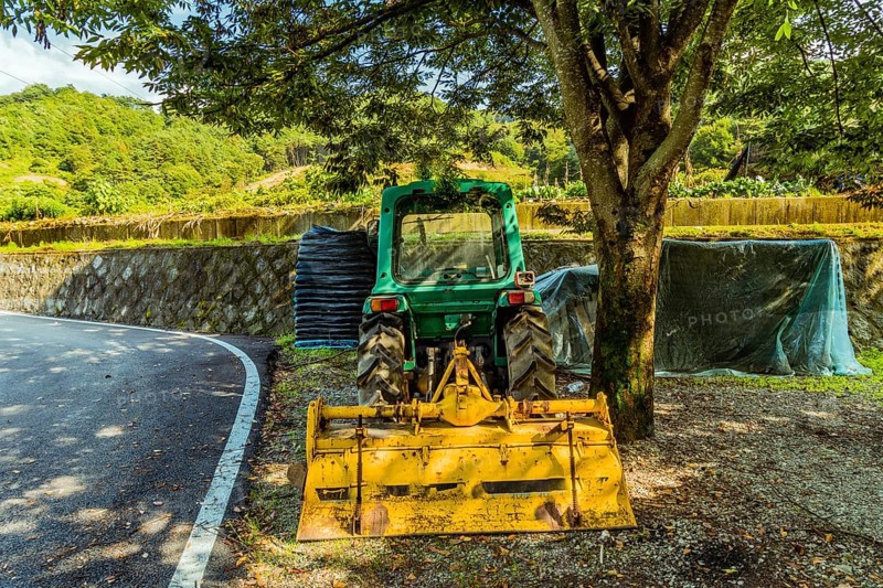 Green tractor with yellow cutting attachment parked under a large shade tree next to a paved road in South Korea