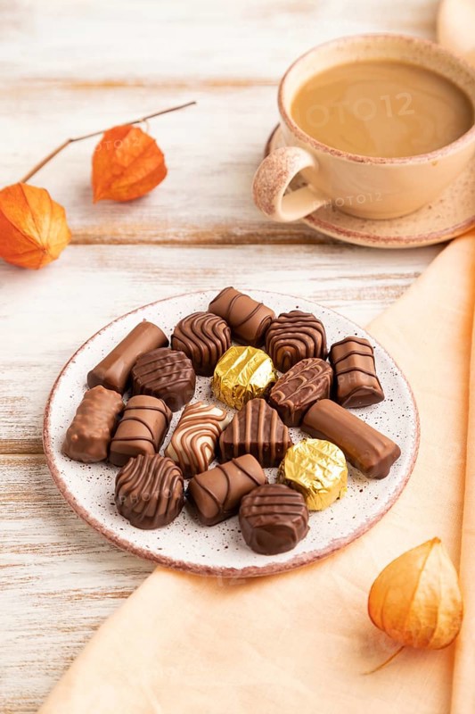 Chocolate candies with cup of coffee and physalis flowers on a white wooden background and orange textile. side view, close up