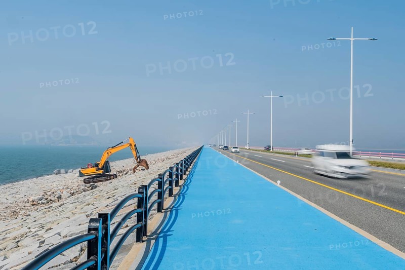 Backhoe sitting on ocean causeway next to blue concrete sidewalk on divided highway with speeding cars passing by. Motion blur caused by slow shutter speed