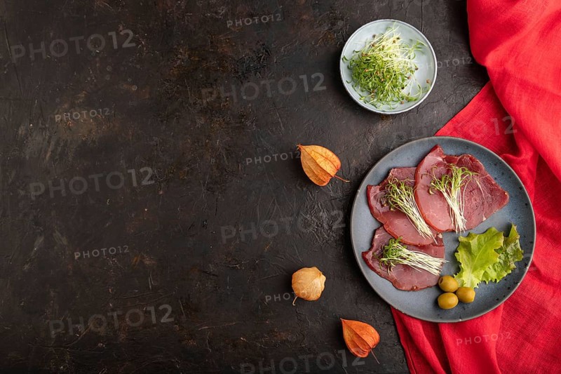 Slices of smoked salted meat with cilantro microgreen on black concrete background and red textile. Top view, flat lay, copy space