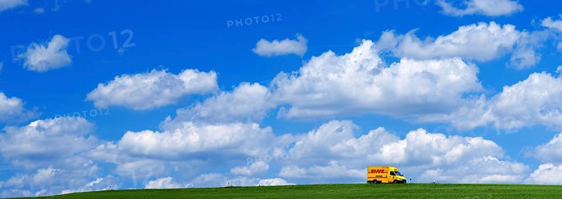 A DHL delivery van on a country road in the middle of meadows with blue sky and many white fair weather clouds, panoramic picture