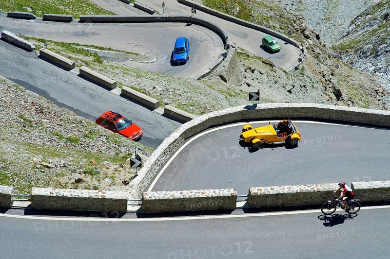 A red, green and blue car, a yellow roadster and a bicycle driver in hairpin curves on the Stelvio Pass, South Tyrol, Italy, Europe