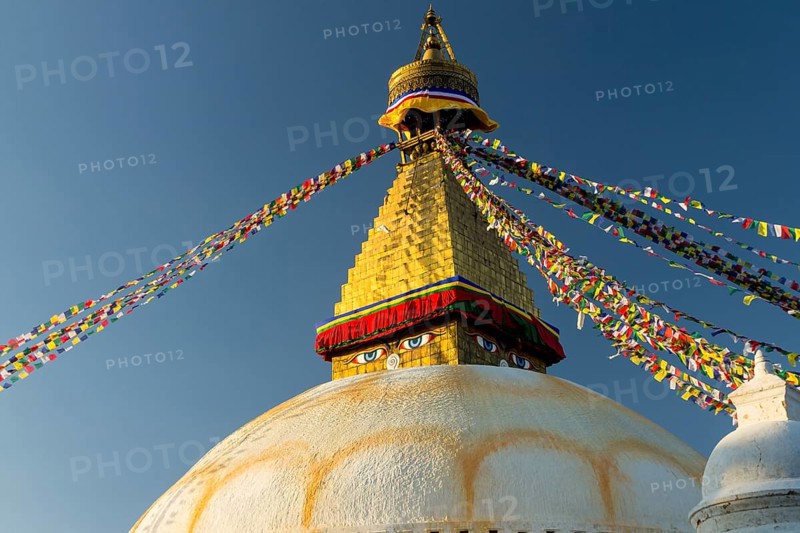 Dome and golden tower of Boudhanath, the Bouddha Stupa, the largest stupa in Nepal, Buddhist religious complex, UNESCO World Heritage Site and one of the main Kathmandu Valley tourist attractions. Seen late in the afternoon on a clear, blue-sky, autumn day in the warm light of setting sun. Eyes depicted on the spire represent the all-seeing wisdom eyes of the Buddha. Buddhist prayer flags hang stretched from the top of the spire. Photographed before the 2015 earthquake. Kathmandu, Bagmati Province, Nepal, Asia