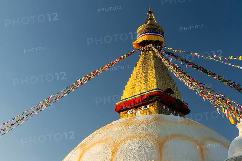 Dome and golden tower of Boudhanath, the Bouddha Stupa, the largest stupa in Nepal, Buddhist religious complex, UNESCO World Heritage Site and one of the main Kathmandu Valley tourist attractions. Seen late in the afternoon on a clear, blue-sky, autumn day in the warm light of setting sun. Eyes depicted on the spire represent the all-seeing wisdom eyes of the Buddha. Buddhist prayer flags hang stretched from the top of the spire. Photographed before the 2015 earthquake. Kathmandu, Bagmati Province, Nepal, Asia