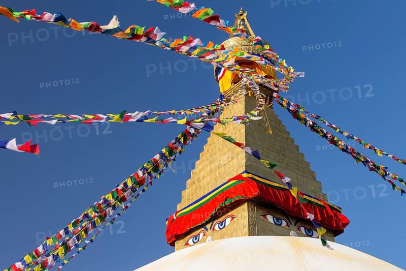 Dome and golden tower of Boudhanath, the Bouddha Stupa, the largest stupa in Nepal, Buddhist religious complex, UNESCO World Heritage Site and one of the main Kathmandu Valley tourist attractions. Seen late in the afternoon on a clear, blue-sky, spring day in the warm light of setting sun. Eyes depicted on the spire represent the all-seeing wisdom eyes of the Buddha. Buddhist prayer flags hang stretched from the top of the spire. Photographed after the 2015 earthquake. Kathmandu, Bagmati Province, Nepal, Asia