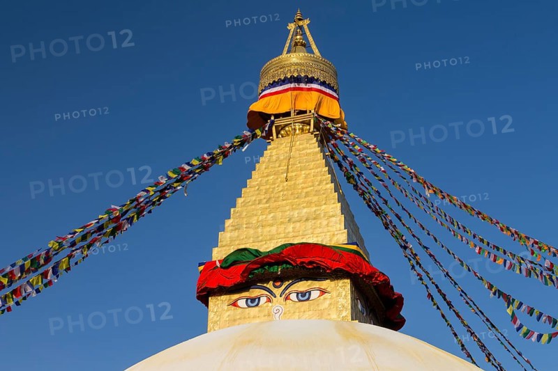 Dome and golden tower of Boudhanath, the Bouddha Stupa, the largest stupa in Nepal, Buddhist religious complex, UNESCO World Heritage Site and one of the main Kathmandu Valley tourist attractions. Seen late in the afternoon on a clear, blue-sky, spring day in the warm light of setting sun. Eyes depicted on the spire represent the all-seeing wisdom eyes of the Buddha. Buddhist prayer flags hang stretched from the top of the spire. Site was rebuilt after the 2015 earthquake. Kathmandu, Bagmati Province, Nepal, Asia