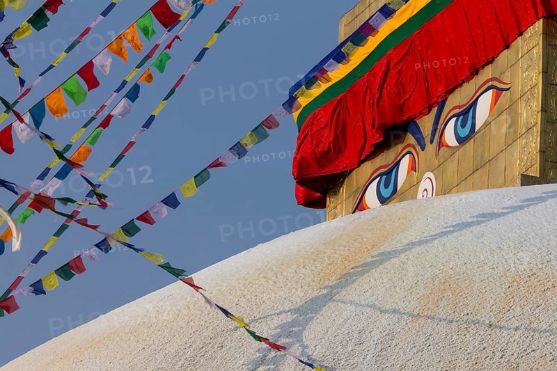 Buddhist prayer flags and eyes of Buddha, the Wisdom Eyes, painted on the golden spire of Boudhanath, the Bouddha Stupa, the largest stupa in Nepal, Buddhist religious complex, UNESCO World Heritage Site and one of the Kathmandu Valley tourist attractions. Seen in the afternoon, on a clear, blue-sky, spring day, when the monument was lit by the setting sun. Photographed after the major reconstruction, that followed the 2015 earthquake, when the site was severely damaged. Kathmandu, Bagmati Province, Nepal, Asia