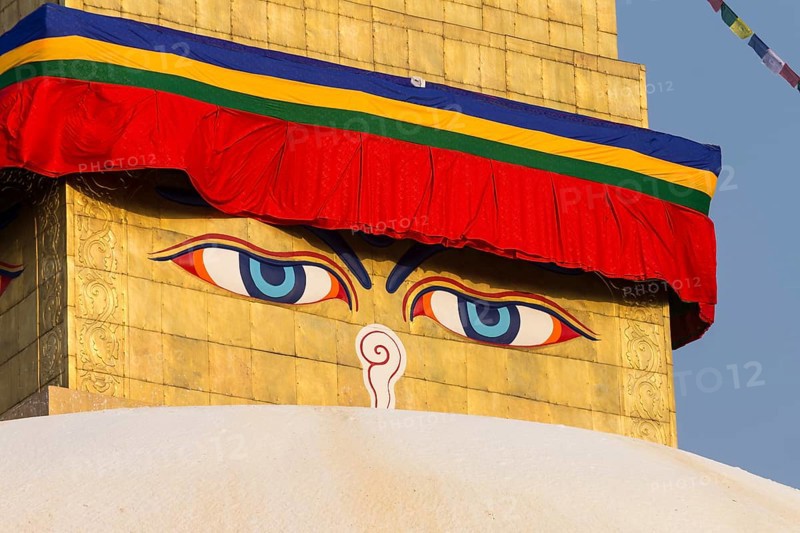 All-seeing eyes of Buddha, the Wisdom Eyes or Buddha's eyes, painted on the golden spire of Boudhanath, the Bouddha Stupa, the largest stupa in Nepal, Buddhist religious complex, UNESCO World Heritage Site and one of the Kathmandu Valley tourist attractions. Seen in the afternoon, on a clear, blue-sky, spring day, when the monument was lit by the setting sun. Photographed after the major reconstruction, that followed the 2015 earthquake, when the site was severely damaged. Kathmandu, Bagmati Province, Nepal, Asia