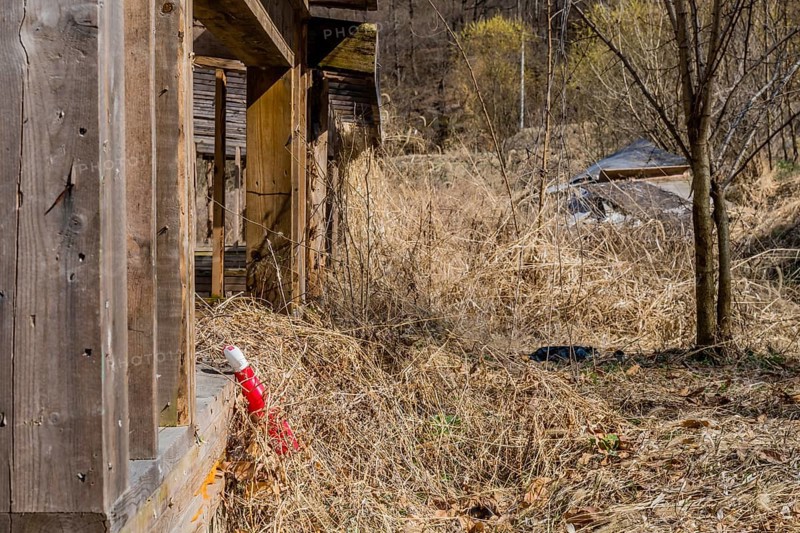Red plastic bottle in dry weeds resting against wooden sill of abandoned building