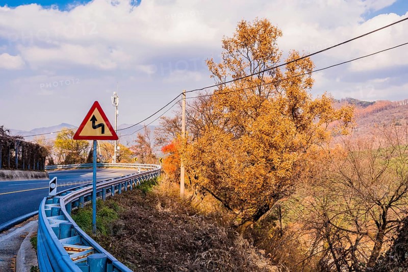 Warning sign next to guardrail on curvy mountain road with cloudy sky in background in South Korea