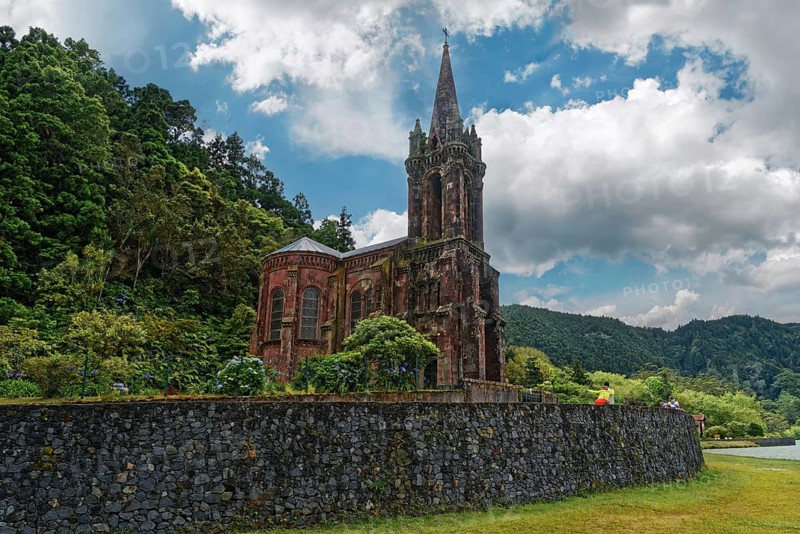 Old gothic church Capela de Nossa Senhora das Vitorias surrounded by trees and a stone wall against a dramatic cloudy sky, Furnas Lake, Furnas, Sao Miguel, Azores, Portugal, Europe