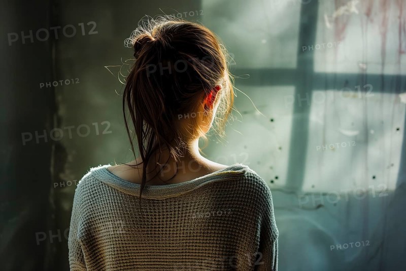 Back view of a young woman shrouded in subdued sunlight near a window, symbolising depression and abuse, AI generated, AI generated