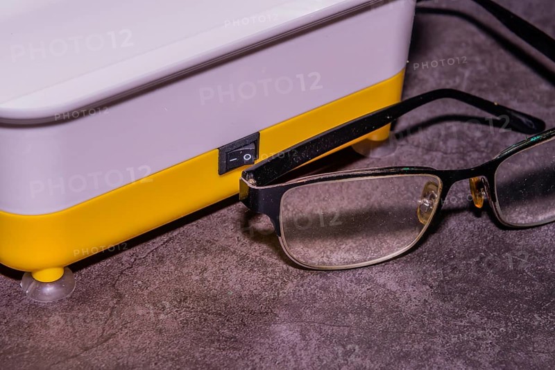 Closeup of pair of wire framed glasses in front of ultrasonic cleaner machine on black textured background