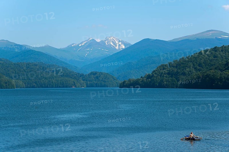 A small boat on a calm blue reservoir with snow-capped mountains in the background, Vidraru Reservoir, Transfogarasan High Road, Transfagarasan, TransfagaraÈ™an, FagaraÈ™ Mountains, Fagaras, Transylvania, Transylvania, Transylvania, Ardeal, Transilvania, Carpathians, Romania, Europe