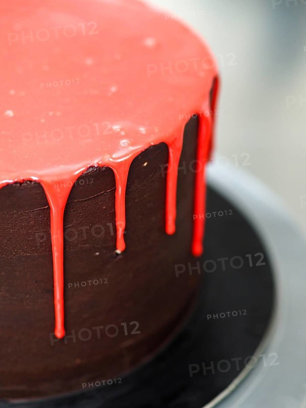 A dark chocolate frosted cake with glossy red dripping icing on a stand