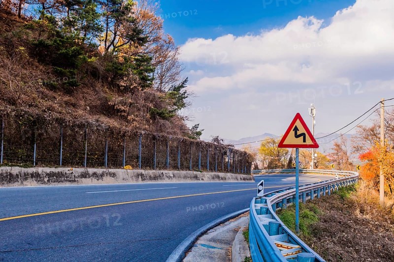 Warning sign next to guardrail on curvy mountain road with cloudy sky in background in South Korea