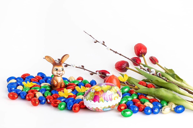 A collection of colourful chocolate eggs and an Easter bunny next to tulips and palm catkins, white background